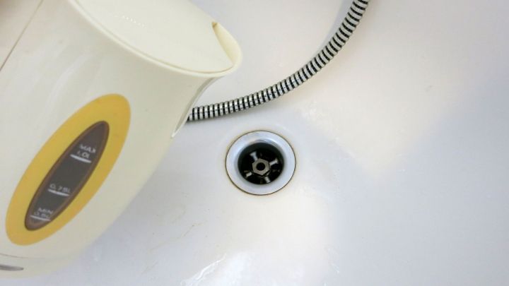 Ein Wasserkocher gie&szlig;t Wasser in ein Abflussbecken. Ein gestreifter Schlauch liegt daneben im wei&szlig;en Waschbecken.