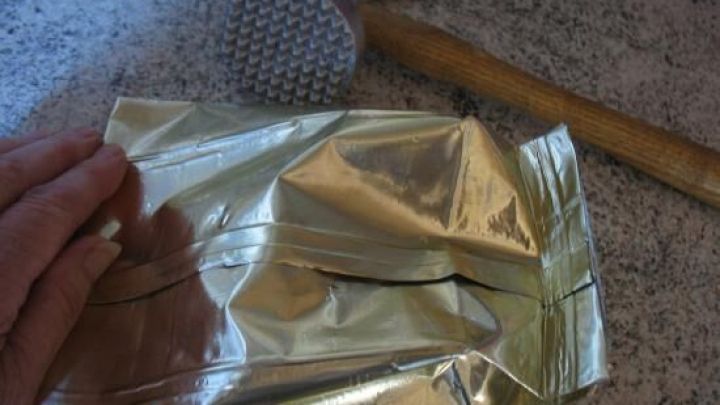 Eine Hand h&auml;lt eine goldene Verpackung, w&auml;hrend ein Hammer bereitliegt, um N&uuml;sse oder Schokolade zu hacken.