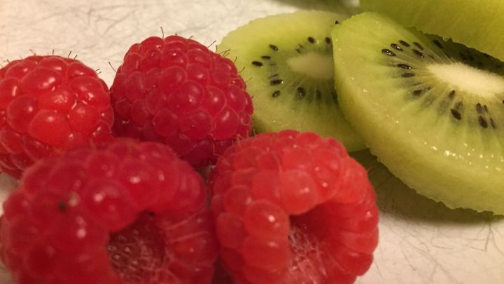 Frische Himbeeren liegen neben d&uuml;nn geschnittenen Kiwis auf einer hellen Arbeitsfl&auml;che, bereit zur Verarbeitung f&uuml;r Himbeerso&szlig;e.
