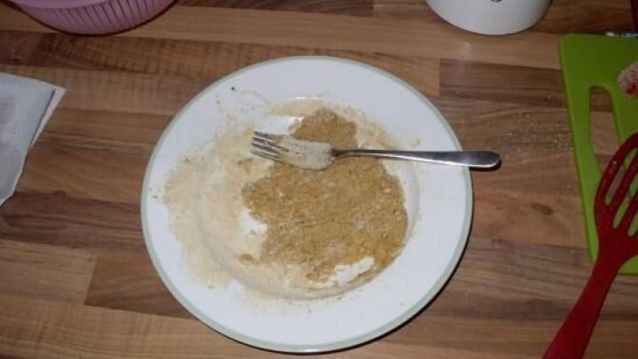 Ein Teller mit Panade und Gabel liegt auf Holzarbeitsplatte. Daneben ein Schneebesen und ein Schneidebrett in der K&uuml;che.