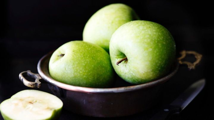 Gr&uuml;ne &Auml;pfel liegen in einer dunklen Sch&uuml;ssel, w&auml;hrend ein halbierter Apfel und ein Messer daneben liegen.
