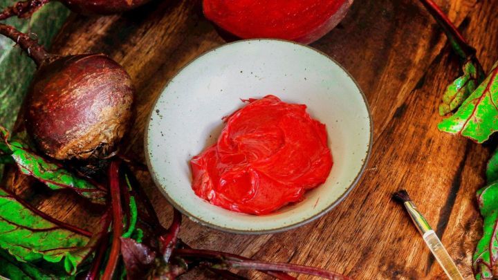 Rote Lippenpflege liegt in einer Sch&uuml;ssel, umgeben von frischen R&uuml;ben und gr&uuml;nen Bl&auml;ttern auf einem Holzbrett.