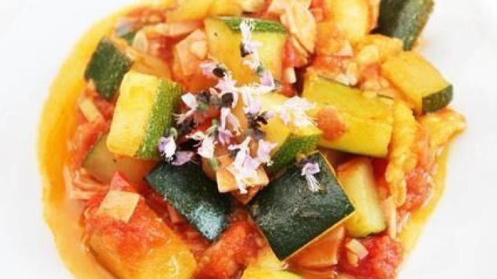 Zucchiniw&uuml;rfel liegen mit Tomaten und Kr&auml;utern garniert auf einem wei&szlig;en Teller, umgeben von hellen Bl&uuml;ten.