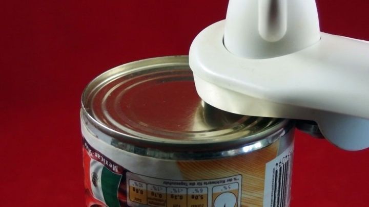 Eine Dosenlasche wird mit einem Dosen&ouml;ffner auf einer Konservendose in einem roten Umfeld ge&ouml;ffnet.