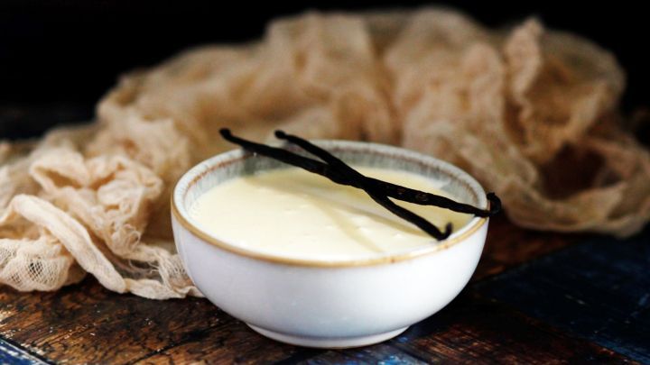 Eine Sch&uuml;ssel mit cremiger Vanilleso&szlig;e liegt auf einem rustikalen Holztisch, dekoriert mit Vanilleschoten und einem sanften Stoff.