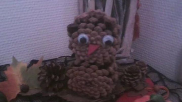 Eine Eulen-Deko aus Tannenzapfen sitzt zwischen Herbstbl&auml;ttern und einer K&uuml;rbisfigur auf einem Tisch vor Sonnenblumen.