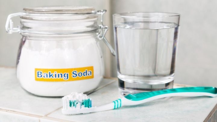 Ein Glas Wasser steht neben einem Beh&auml;lter mit Speisenatron und einer Zahnb&uuml;rste auf einer hellen Oberfl&auml;che.