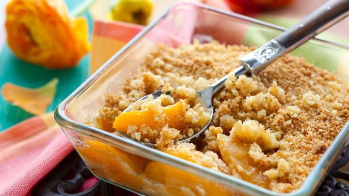 Ein Pfirsich-Crumble in einer Glassch&uuml;ssel wird mit einem L&ouml;ffel serviert, umgeben von bunten Tischdecken und Blumen.