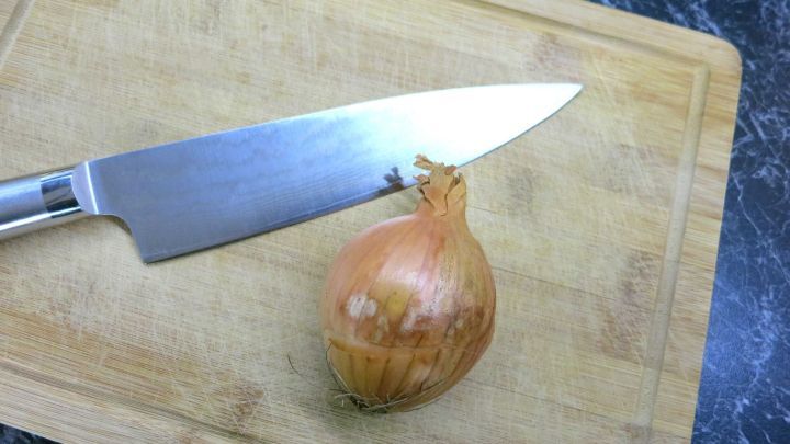 Eine Zwiebel liegt auf einem Holzbrett neben einem scharfen K&uuml;chenmesser, bereit zum Schneiden in einer K&uuml;chenumgebung.