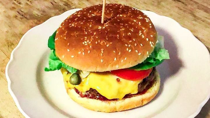 Ein saftiger Burger mit K&auml;se und frischem Gem&uuml;se liegt auf einem wei&szlig;en Teller auf einem Holztisch.