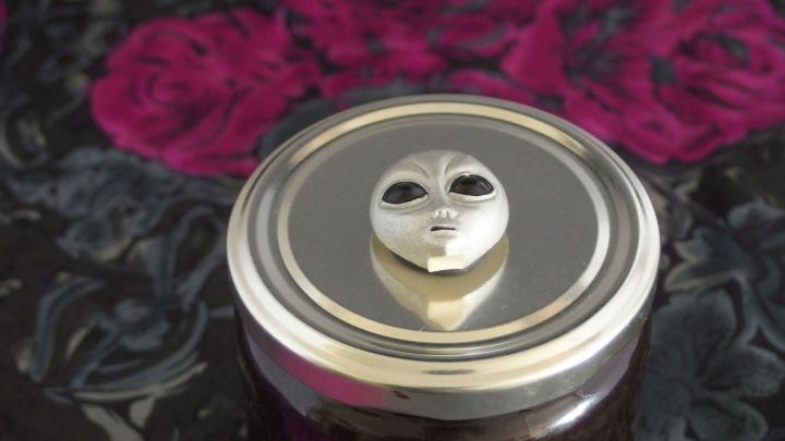 Ein Marmeladenglas mit einem kunstvollen Deckel, der eine stilisierte Figur zeigt, steht auf einem blumigen Hintergrund.