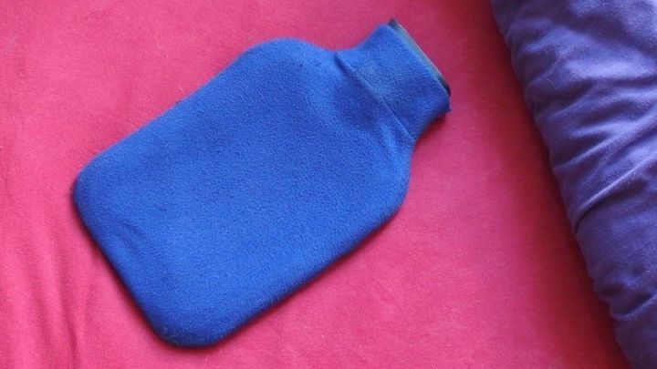 Eine blaue W&auml;rmflasche liegt auf einem roten Bettlaken, bereit daf&uuml;r, w&auml;hrend der kalten N&auml;chte im Herbst und Winter W&auml;rme zu spenden.