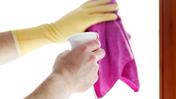Eine Hand mit gelben Gummihandschuhen h&auml;lt einen Reinigungsspray und wischt mit einem pinken Tuch ein Fenster.