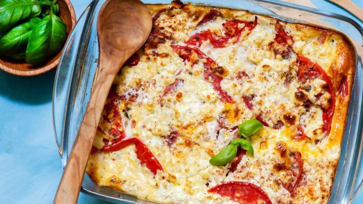 Ein Auflauf aus mediterranem Schafsk&auml;se und Gem&uuml;se liegt in einer Glasform, umgeben von frischem Basilikum und Holzutensilien.