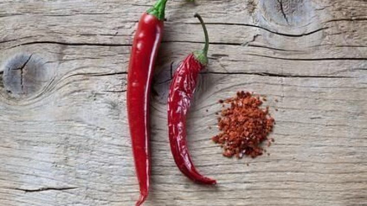 Peperoni liegen auf rustikalem Holz, eine ist ganz, die andere getrocknet und zu Pulver gemahlen, daneben kleine H&auml;ufchen.