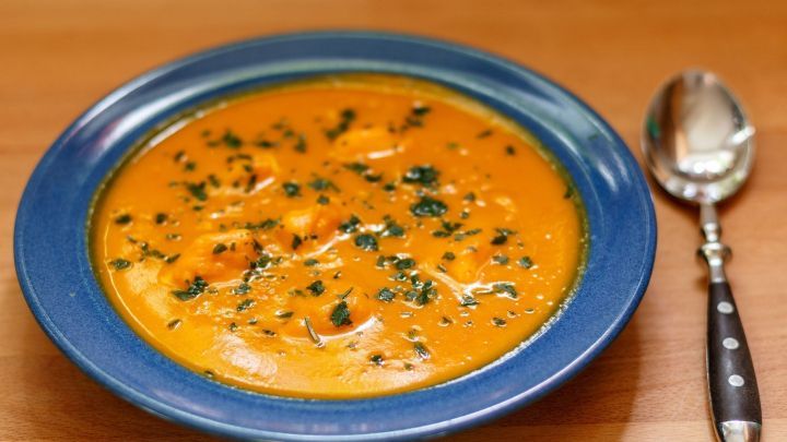 Eine orange K&uuml;rbiscremesuppe steht in einer blauen Schale, dekoriert mit Kr&auml;utern, neben einem L&ouml;ffel auf einem Holztisch.
