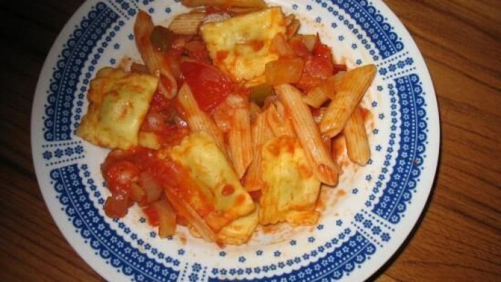 Eine Nudelpfanne mit Penne und Ravioli liegt in einem blau-wei&szlig; gemusterten Teller auf einem Holztisch. Text: "Mit Ravioli".