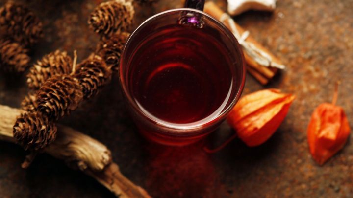 Ein Glas Gl&uuml;hwein steht auf einer rustikalen Oberfl&auml;che, umgeben von Zimtstangen und dekorativen Leckereien.