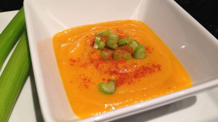 Eine cremige K&uuml;rbissuppe mit Kokosmilch und Kreuzk&uuml;mmel ist in einer wei&szlig;en Schale angerichtet, garniert mit gr&uuml;nen Zwiebelst&uuml;cken und Paprika. Am Rand liegen frische Selleriestangen.