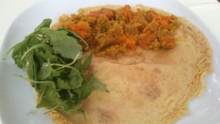 Ein gro&szlig;er, d&uuml;nner Pfannkuchen liegt auf einem wei&szlig;en Teller, gef&uuml;llt mit K&uuml;rbis und roten Linsen, umgeben von frischem Salat.