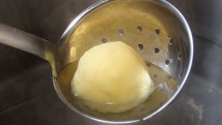 Ein Kartoffelkn&ouml;del liegt in einem Siebl&ouml;ffel, w&auml;hrend er in einem Topf mit kochendem Wasser gegart wird.
