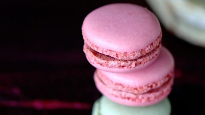 Bunte Macarons stehen auf einem Holzuntergrund, w&auml;hrend im Hintergrund eine Sch&uuml;ssel sichtbar ist.