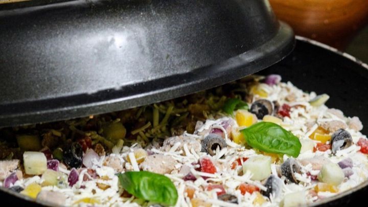 Eine Pfanne mit einer Pizza, die mit frischem Gem&uuml;se und K&auml;se belegt ist, wird mit einem Deckel abgedeckt.