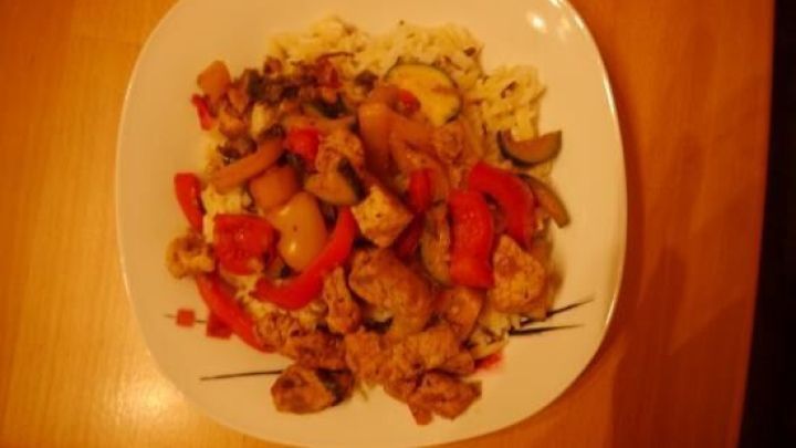 Eine bunte Gyrospfanne mit Fleisch, Paprika und Gem&uuml;se auf einem wei&szlig;en Teller, serviert auf einem Holzuntergrund.