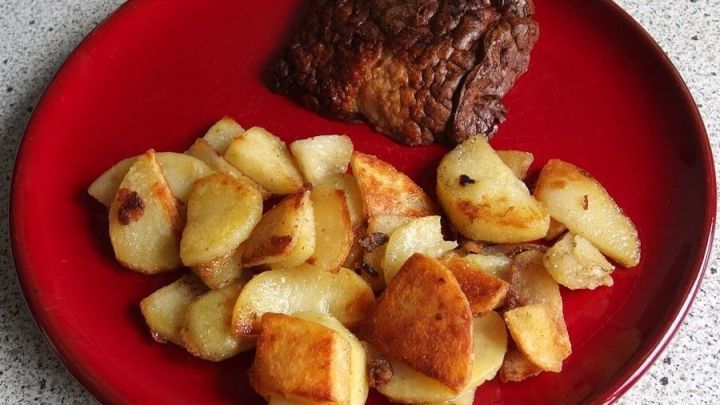 Bratkartoffeln liegen gebraten auf einem roten Teller neben einem St&uuml;ck Fleisch, in einer gem&uuml;tlichen K&uuml;chenszene.