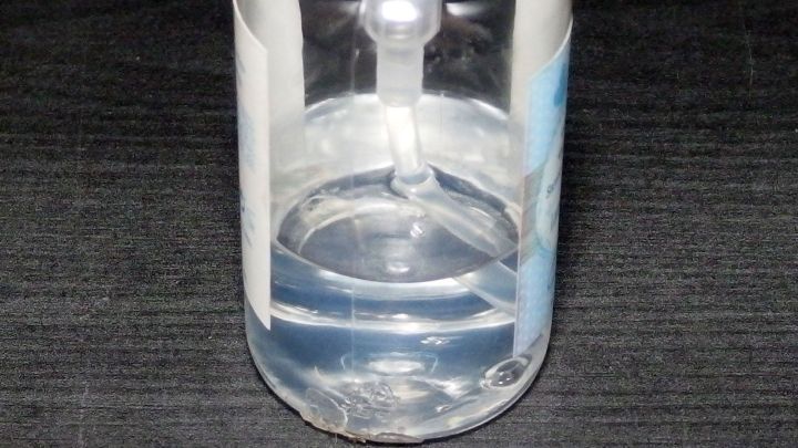 Eine transparente Flasche enth&auml;lt fl&uuml;ssige Handseife und steht auf einer dunklen Oberfl&auml;che in einem neutralen Umfeld.