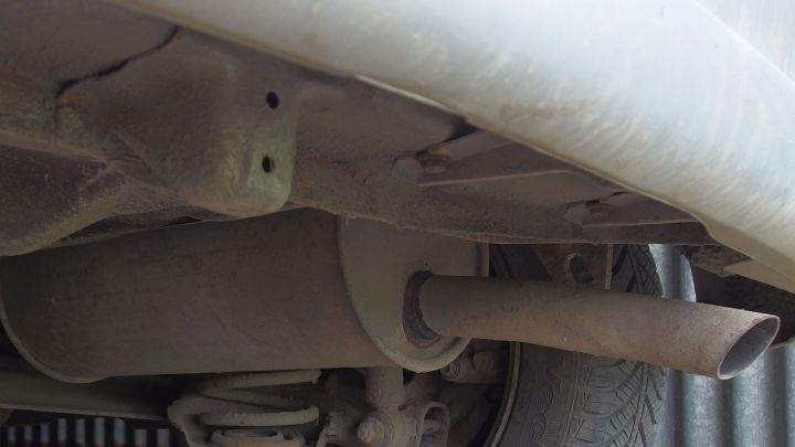 Ein Auspuff h&auml;ngt unter einem Fahrzeug, w&auml;hrend Abgase austreten und sich in einer grauen Umgebung verbreiten.