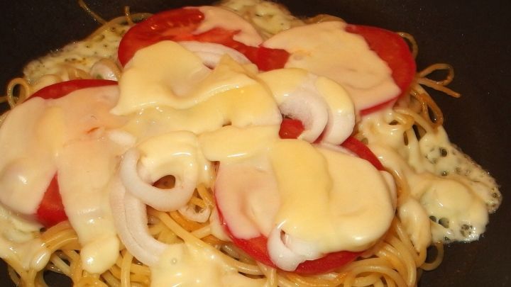 Eine Pfanne mit gebratenen Spaghetti, belegt mit Tomatenscheiben und geschmolzenem K&auml;se, dampft in der K&uuml;che.