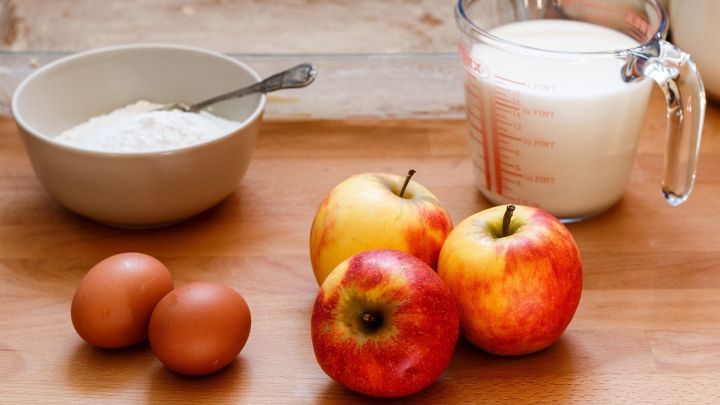 Auf einem Holzbrett liegen &Auml;pfel, Eier und eine Sch&uuml;ssel mit Mehl, w&auml;hrend ein Messbecher Milch bereitsteht.