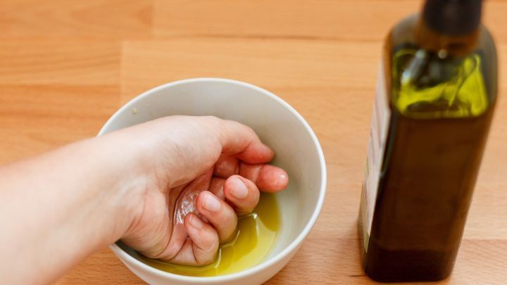Eine Hand mit rissigen Fingern taucht in eine Sch&uuml;ssel mit &Ouml;l, w&auml;hrend eine Flasche Oliven&ouml;l daneben steht.