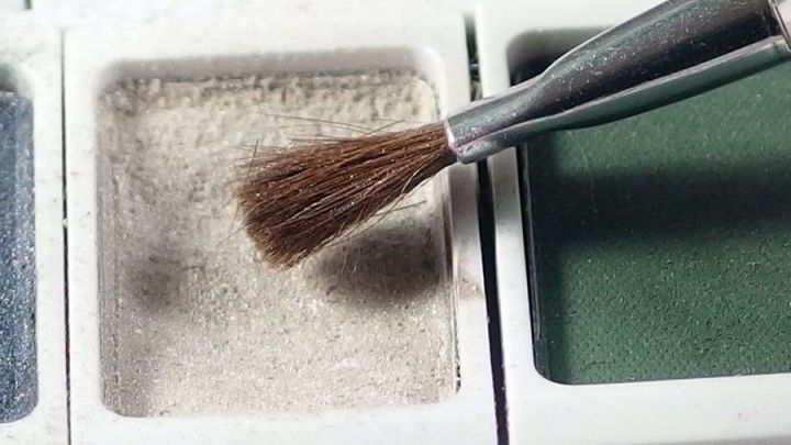 Ein Pinsel hebt einen gl&auml;nzenden Lidschatten in einer Palette an, um ihn gleich aufzutragen, um l&auml;ngerhaltende Farbe zu zeigen.