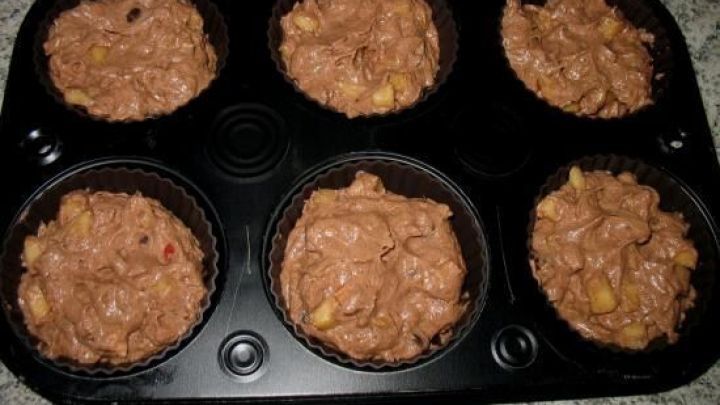 In einer Muffinform liegen sechs ungebackene Muffins mit Schokoladen-Apfel-Teig, bereit zum Backen in einer hellen K&uuml;che.