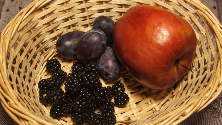 Eine rot gl&auml;nzende Apfel sitzt neben blauen Pflaumen und schwarzen Brombeeren in einem geflochtenen Korb.
