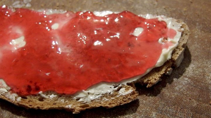 Ein Brotst&uuml;ck mit Frischk&auml;se und rotem Gelee liegt auf einem Holztisch, bereit zum Verzehr als Geschmackserlebnis.