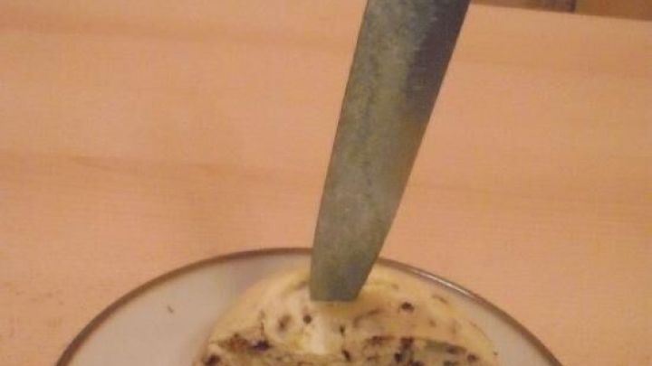 Ein St&uuml;ck warmer Kuchen auf einem Teller; ein Messer steckt darin. Der Tisch und ein gemusterter Stuhl sind im Hintergrund.