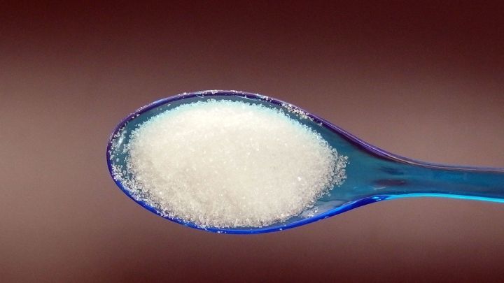 Ein blauer Teel&ouml;ffel mit Zucker steht im Vordergrund, umgeben von einem sanften, braunen Hintergrund.