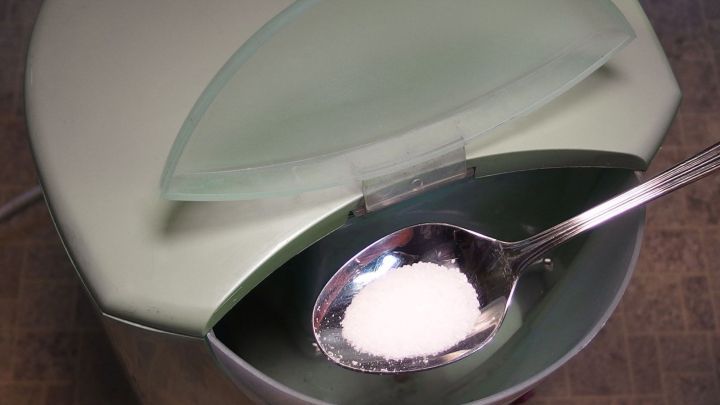 Eine Kaffeemaschine mit einem offenen Deckel, in dem ein Teel&ouml;ffel mit Zitronens&auml;ure liegt, steht auf einer Arbeitsfl&auml;che.