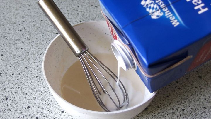 Eine R&uuml;hrsch&uuml;ssel mit einem Schneebesen wird gef&uuml;llt, w&auml;hrend ein Karton mit Halbfettmilch dar&uuml;ber geleert wird.