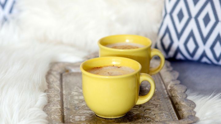 Zwei gelbe Tassen mit Getr&auml;nken stehen auf einem Tablett auf einer weichen Unterlage, umgeben von dekorativen Kissen.