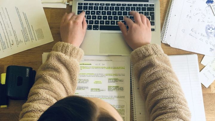 Ein Laptop wird genutzt, w&auml;hrend H&auml;nde auf der Tastatur tippen. Der Tisch ist umgeben von Notizen und Unterlagen.