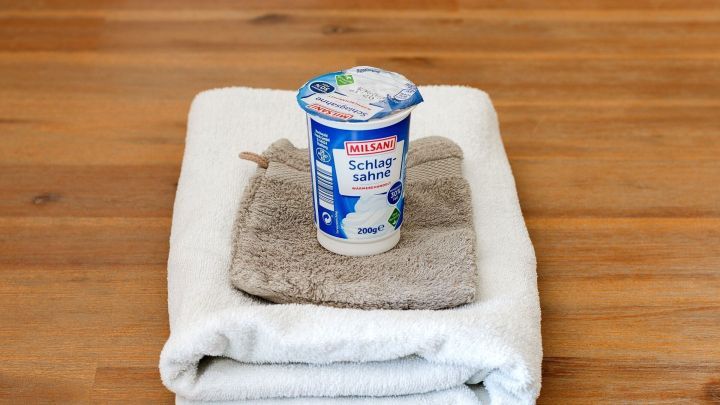 Ein Becher Milsani Schlagsahne steht auf einem hellen Handtuch und einem Waschhandschuh auf einem Holztisch.