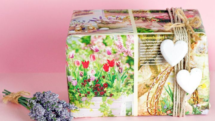 Ein liebevoll verpacktes Geschenk in buntem Zeitungspapier liegt auf einem rosa Hintergrund, umgeben von Lavendel.