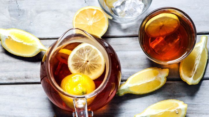 Eine Kanne Zitronen-Eistee steht auf dem Tisch, umgeben von Zitronenscheiben, w&auml;hrend Gl&auml;ser mit Eisf&uuml;llung bereitstehen.