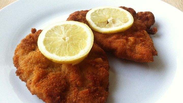 Zwei knusprige Schnitzel auf einem wei&szlig;en Teller, garniert mit Zitronenscheiben, an einem hellen Holzuntergrund.