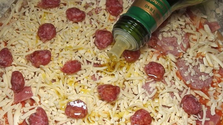 Eine Tiefk&uuml;hlpizza wird mit Oliven&ouml;l betr&auml;ufelt und liegt auf einem Backblech, bereit zum Backen im Ofen.