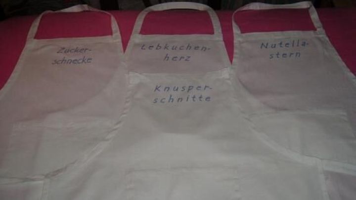 Drei wei&szlig;e Kochsch&uuml;rzen mit handgeschriebenen Namen h&auml;ngen nebeneinander in einer gem&uuml;tlichen K&uuml;chenecke.